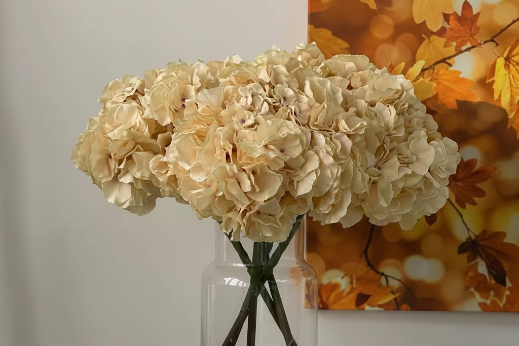 A bouquet of cream-colored artificial hydrangea flowers arranged in a clear glass vase, set against a neutral wall with a framed autumn leaf print in the background.