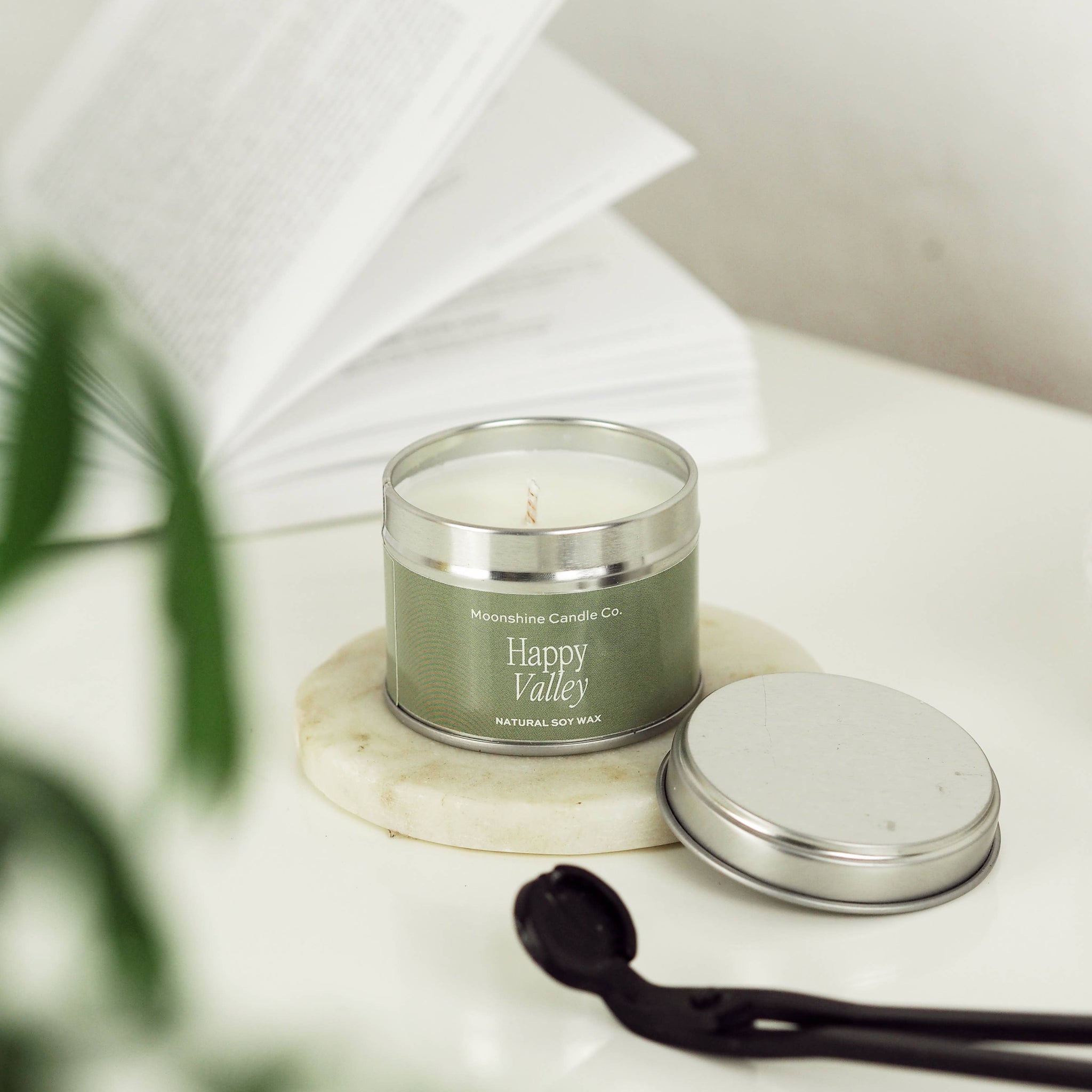 A silver tin candle labeled 'Happy Valley' by Malibu Coast Candle Co sits on a white marble coaster, with its silver lid placed beside it on a white surface. An open book and blurred green leaves are softly in the background.
