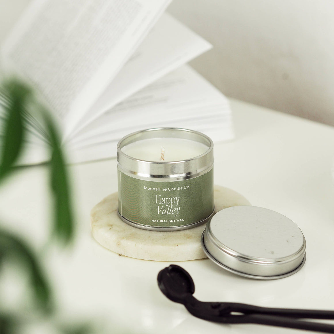 A silver tin candle labeled 'Happy Valley' by Malibu Coast Candle Co sits on a white marble coaster, with its silver lid placed beside it on a white surface. An open book and blurred green leaves are softly in the background.
