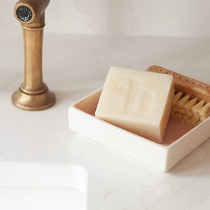 Bar of soap in a dish with a brush on a marble surface next to a brass faucet.