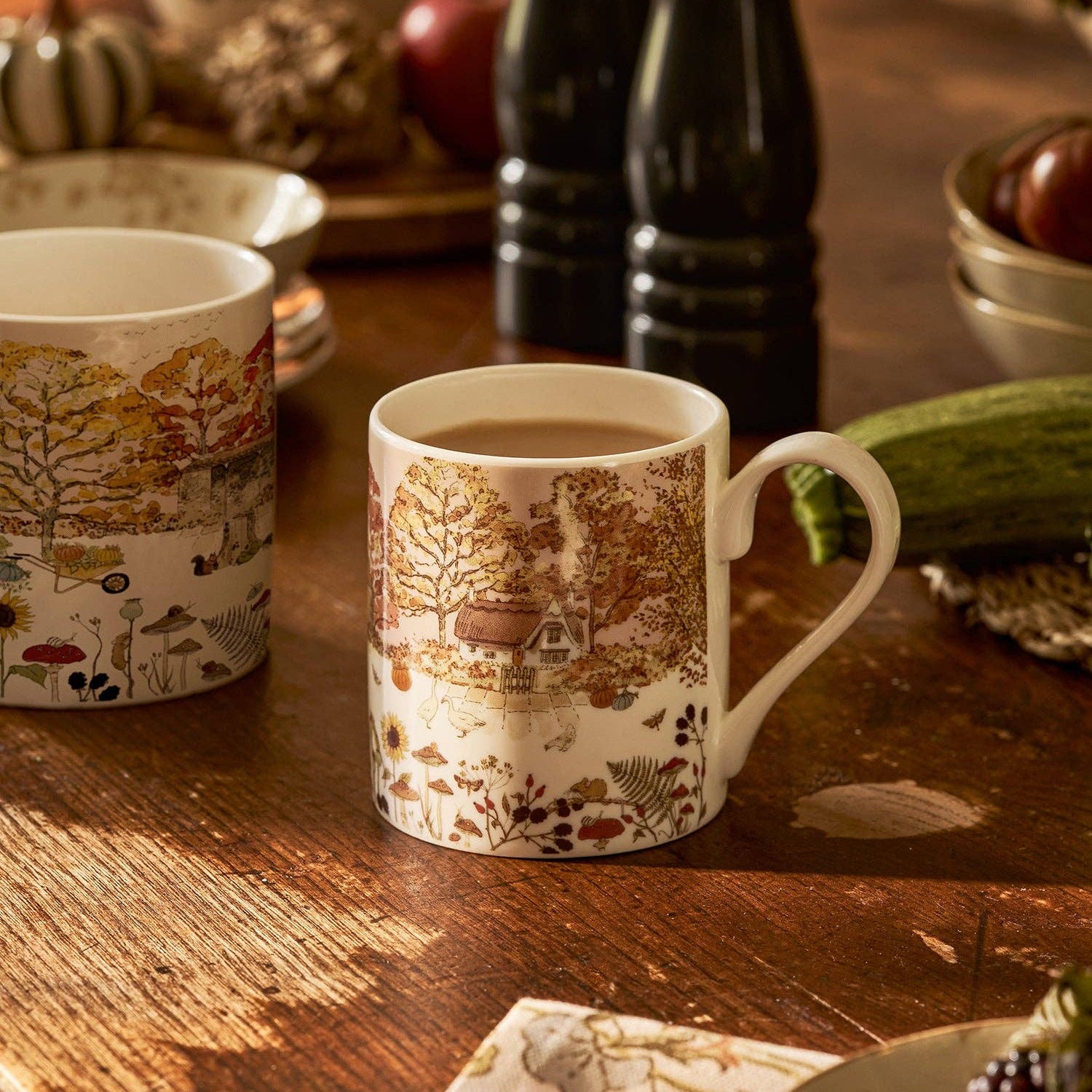 Two ceramic mugs with an autumn forest and cottage landscape print, displayed on a rustic wooden table with seasonal decor in the background.