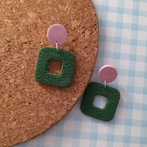 Green square earrings with pink circular tops on a cork board and checkered fabric background
