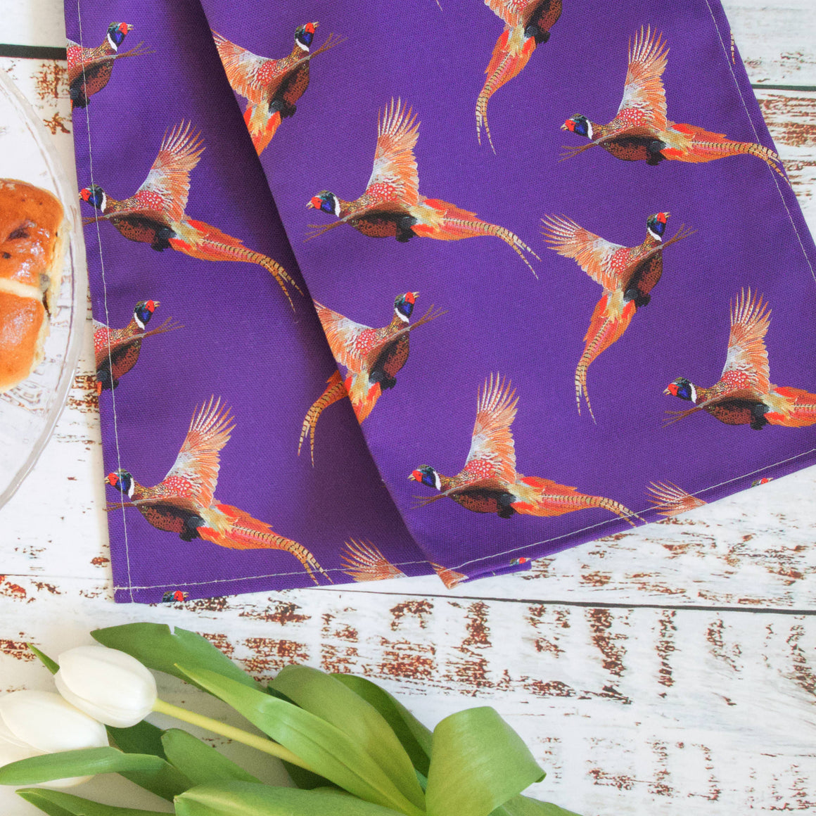 Purple fabric with pheasant pattern on a wooden surface with a plate of food and flowers.