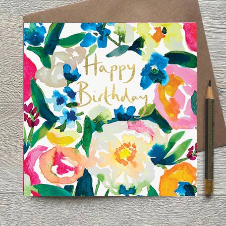 A square floral birthday card with the words 'Happy Birthday' in gold script, placed in front of a brown envelope on a light wood surface. The card features a colorful watercolor floral pattern with blue, pink, yellow, green, and white flowers.