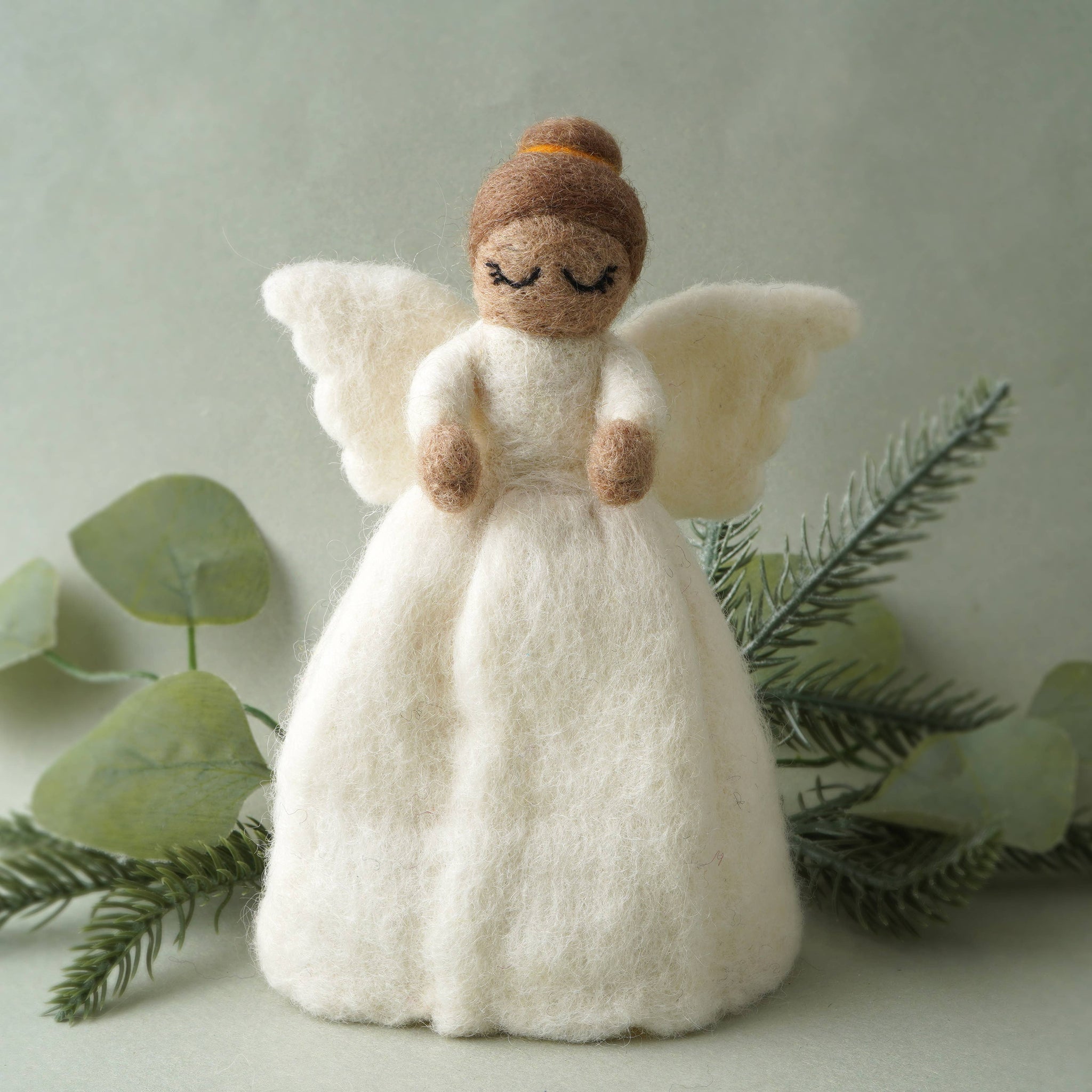 Felted angel figurine with brown hair and white wings, surrounded by greenery on a light background