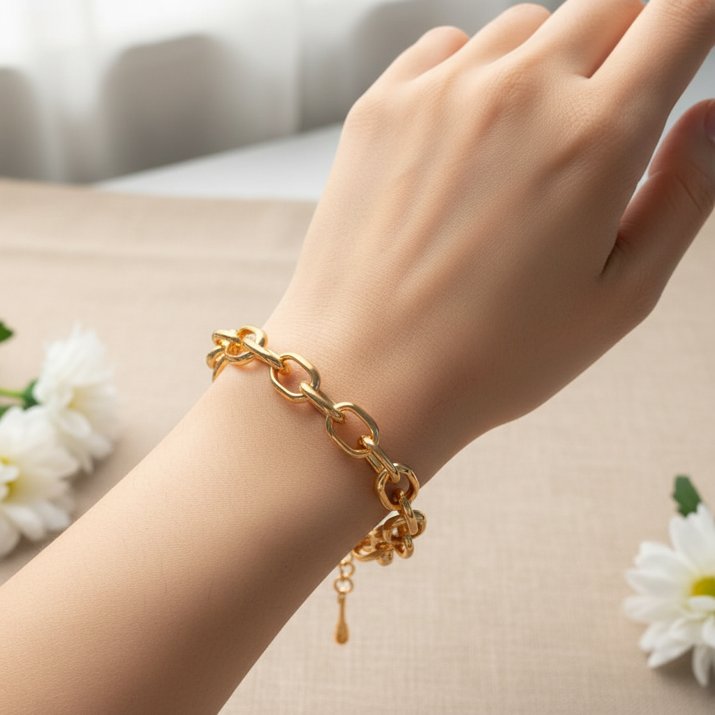 A gold chain link bracelet with a small dangling charm, shown worn on a person's wrist against a soft beige background with white flowers.