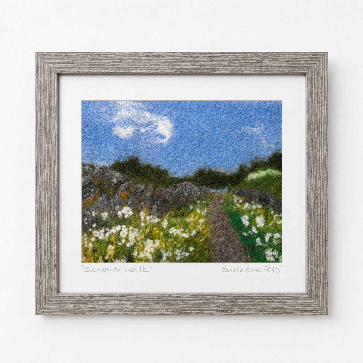 A framed textured art print depicting a summer meadow with a blue sky, white clouds, and wildflowers, set in a rustic gray wooden frame against a white wall.