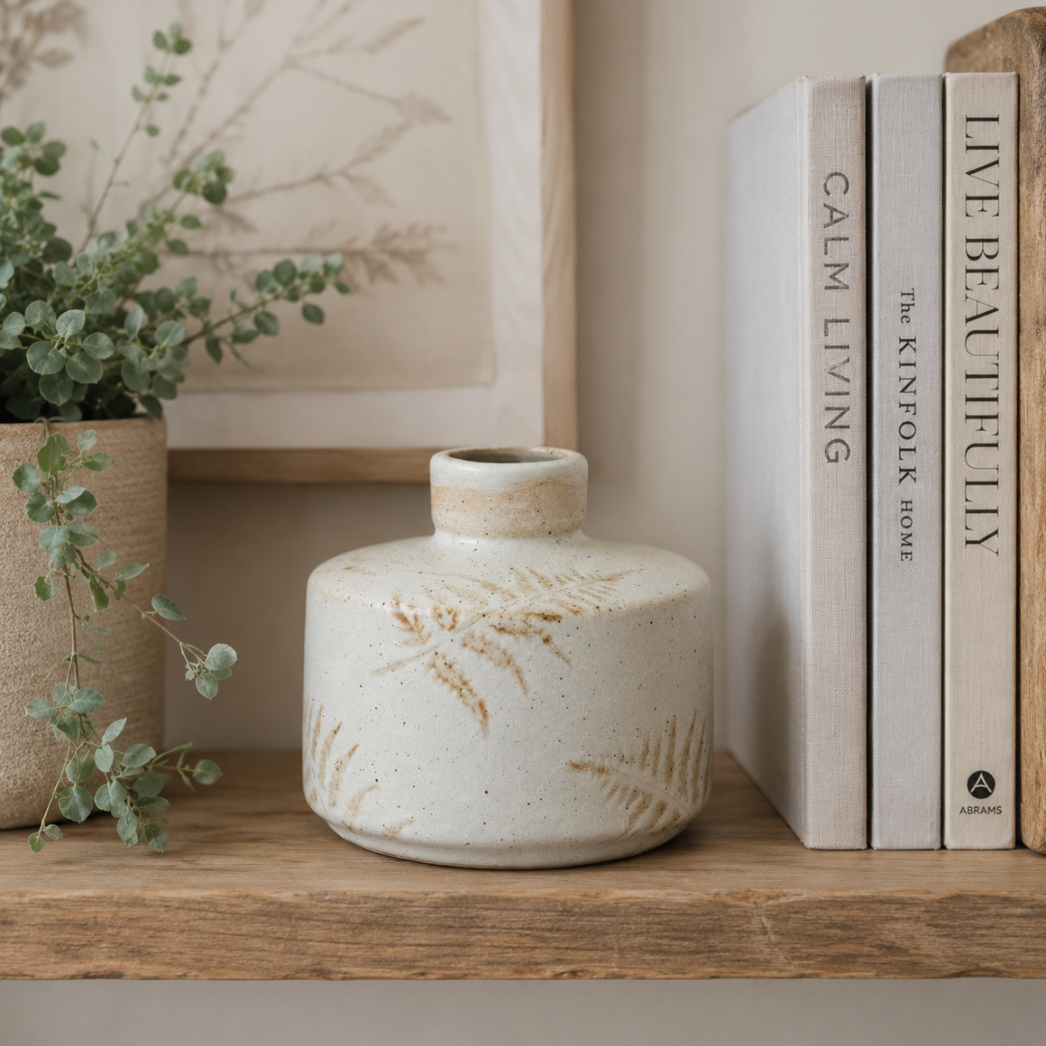 Cream speckled ceramic vase with embossed fern leaf design, styled on a wooden shelf beside greenery and neutral books.