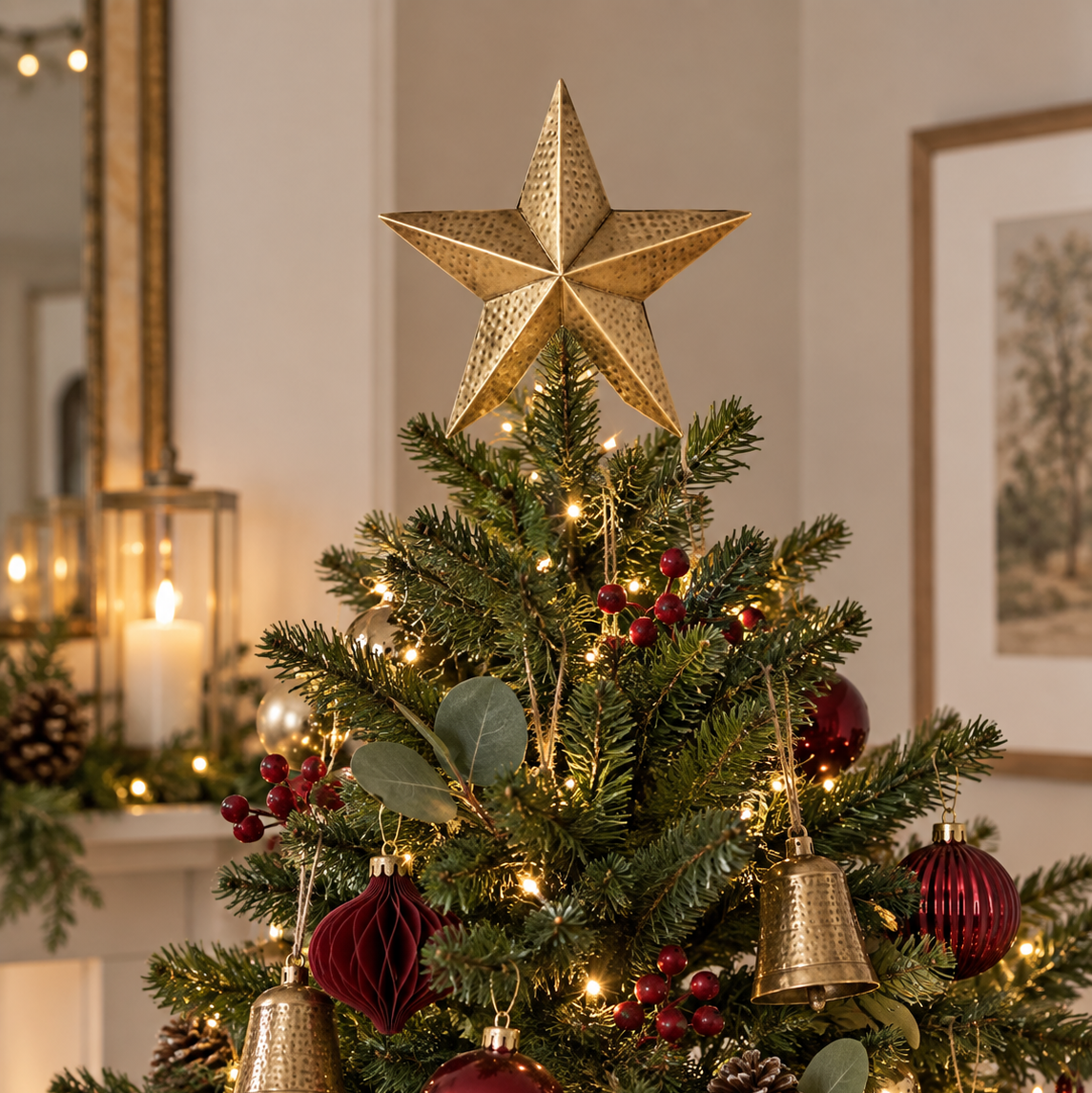 A decorated Christmas tree in a warmly lit indoor setting, topped with a large metallic gold star. The tree is adorned with green branches, warm white string lights, shiny red and gold ornaments, pinecones, and eucalyptus leaves.