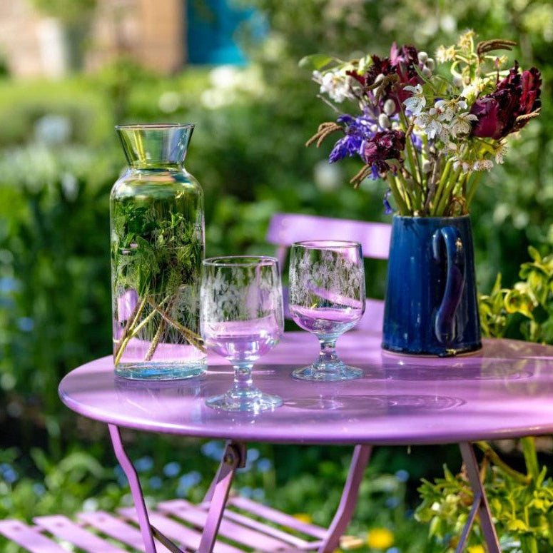 Pink table with a vase, glasses, and flowers in a garden setting