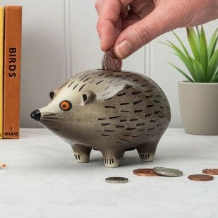 Hedgehog-shaped piggy bank with a hand placing a coin inside, on a light surface with a plant and book in the background.