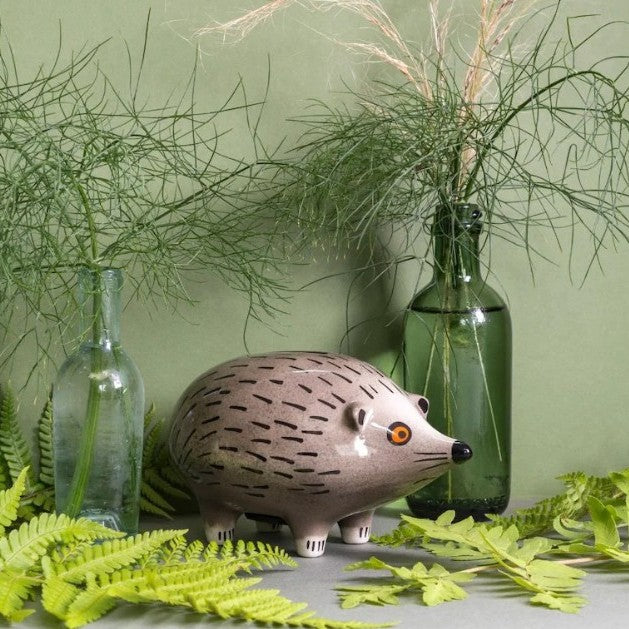 Decorative hedgehog figure surrounded by greenery and bottles on a light surface.