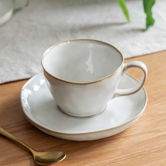 White teacup with gold trim on a matching saucer, placed on a wooden surface with a gold spoon.