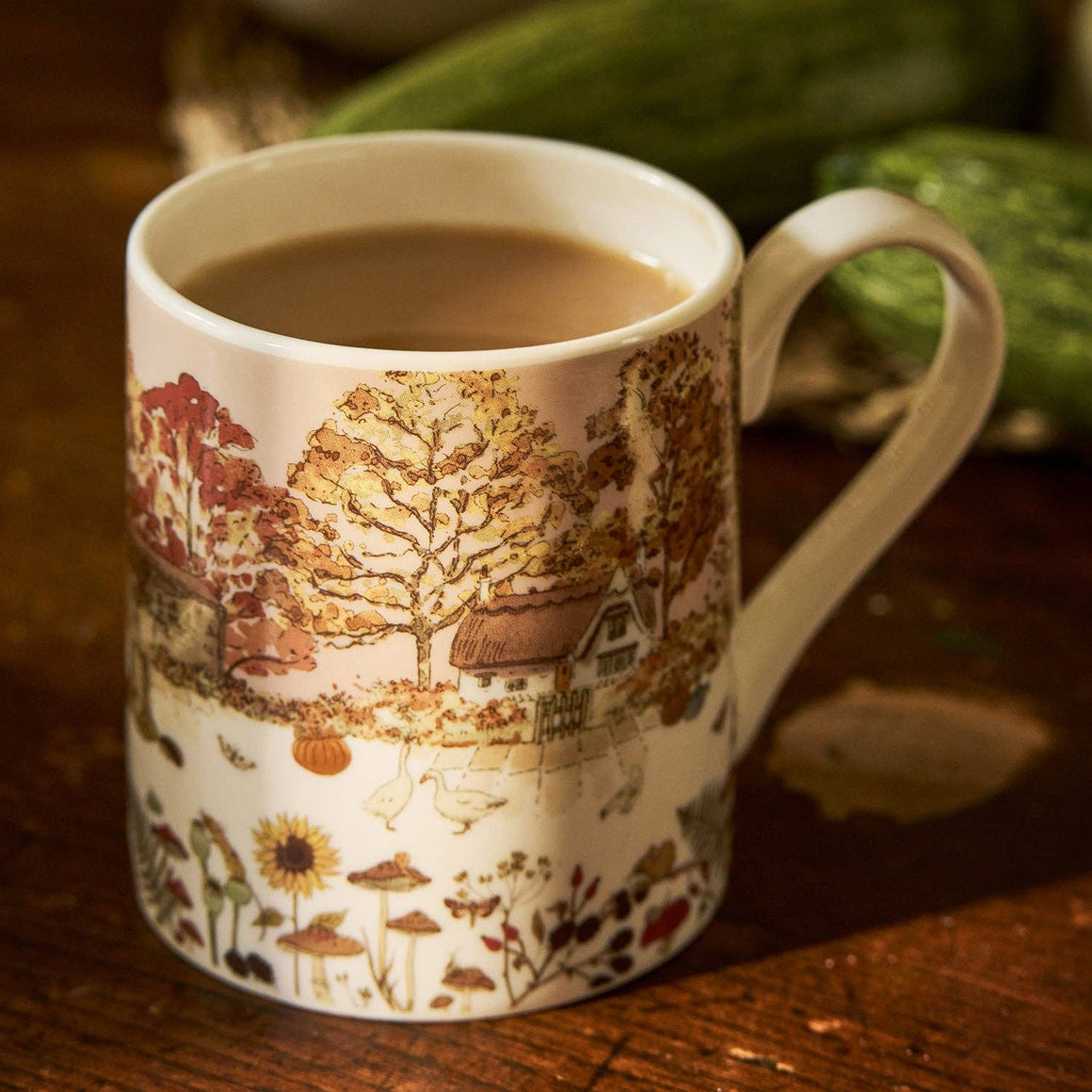 Two ceramic mugs with an autumn forest and cottage landscape print, displayed on a rustic wooden table with seasonal decor in the background.