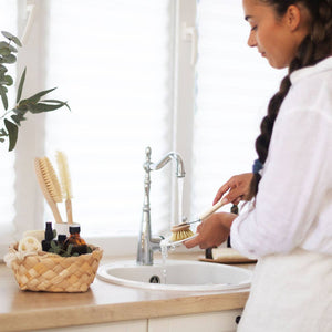 Woman washing dishes in a kitchen with a basket of cleaning supplies on the counter.