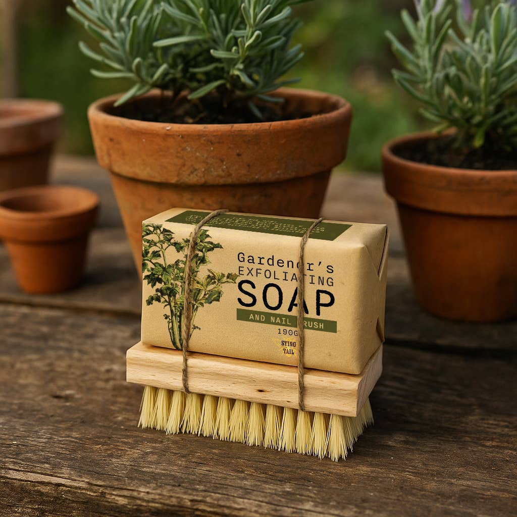 Gardener's Exfoliating Soap and brush set on a wooden surface with potted plants in the background.