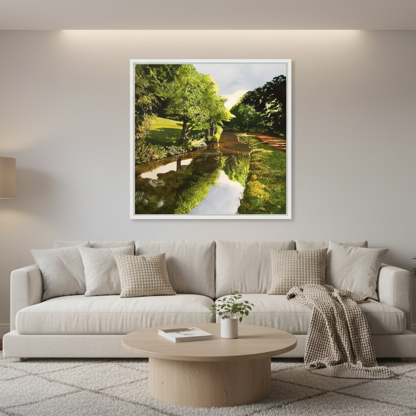 A framed landscape art print depicting a lush green park with a reflective pond, displayed on a light gray wall above a modern beige sofa in a styled living room.