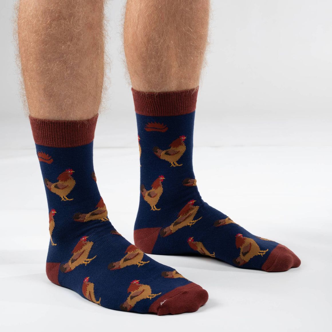 A pair of novelty hen socks with a navy blue body, printed with brown and yellow rooster illustrations, and finished with a dark brown toe cap. They are displayed flat on a light wood grain surface, still attached to their paper packaging.