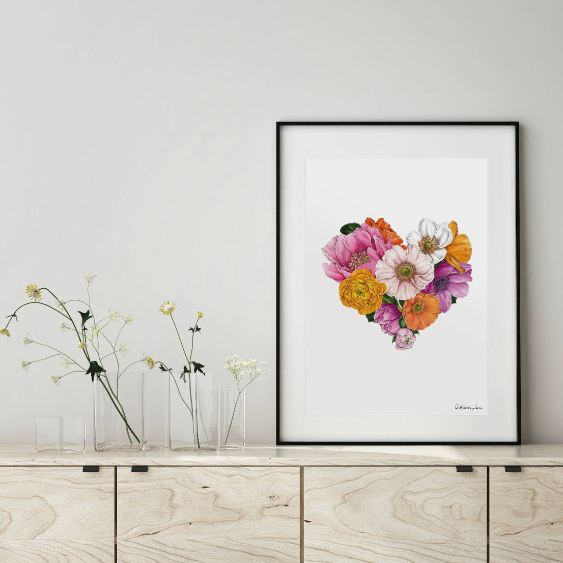 A framed print of a heart-shaped arrangement of colorful flowers displayed on a wooden sideboard.